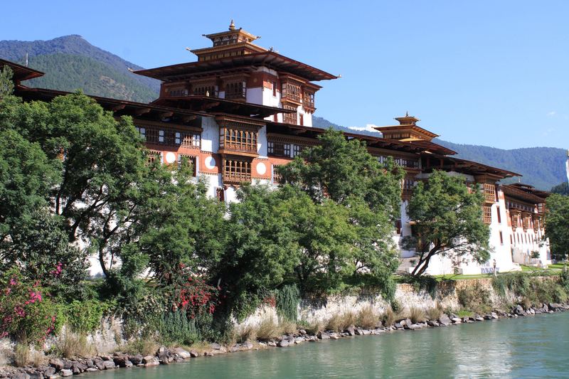 Traditional Bhutanese dzong, a fortress-monastery with carved wooden windows, beside a turquoise river with forested mountains behind