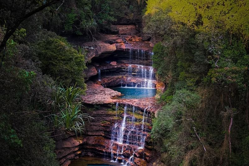 Tiered waterfalls and turquoise pools among red rock ledges and dense green forest in Meghalaya