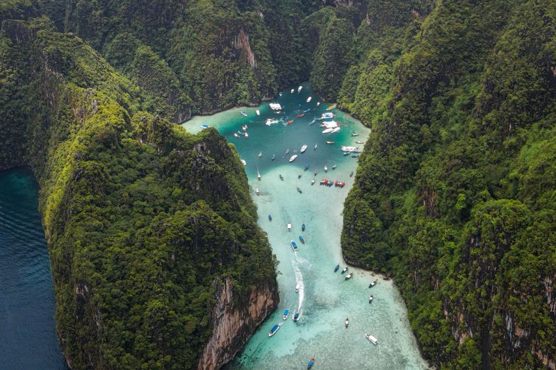Aerial view of a turquoise lagoon walled by steep jungle-covered limestone cliffs, with many longtail boats, in the Phi Phi Islands, Thailand