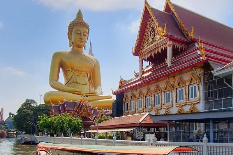 Giant seated golden Buddha statue beside a temple on a canal in Thailand, with a colourful longtail boat in the foreground